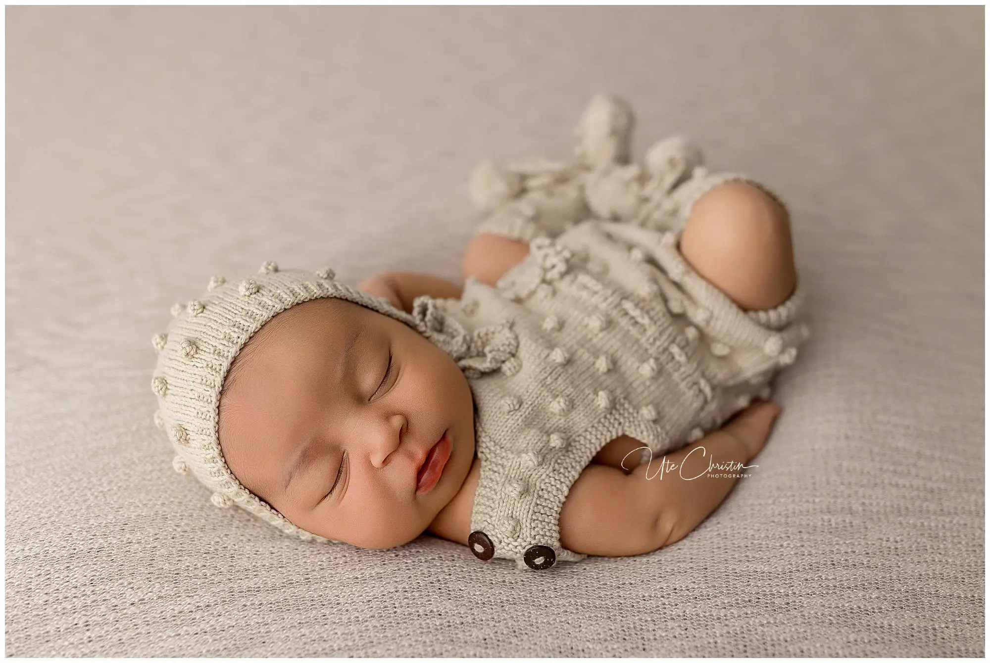 A newborn baby sleeps peacefully on a soft, light-colored blanket, wearing a knitted cream outfit with a matching hat—an adorable moment perfect for families attending Mommy And Me Classes In Milford CT. The baby is curled up with lips slightly parted.