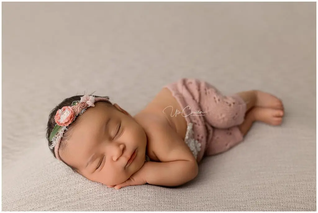 A newborn baby sleeps on their side with a peaceful smile, wearing a pink floral headband and soft pink pants, on a beige blanket—perfect for families seeking memories before starting Mommy And Me Classes In Milford CT.