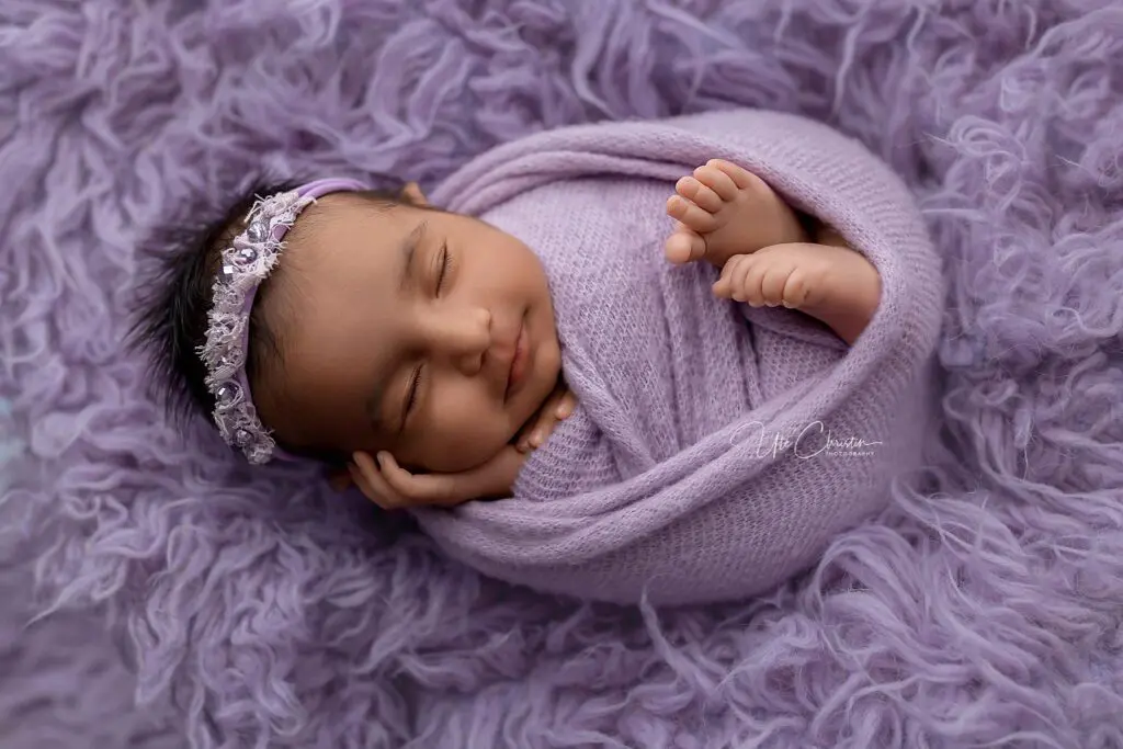 A newborn baby wrapped in a soft lavender blanket, wearing a matching floral headband, sleeps peacefully on a fluffy lavender background—perfect inspiration for those seeking baby shower venues in Milford CT.