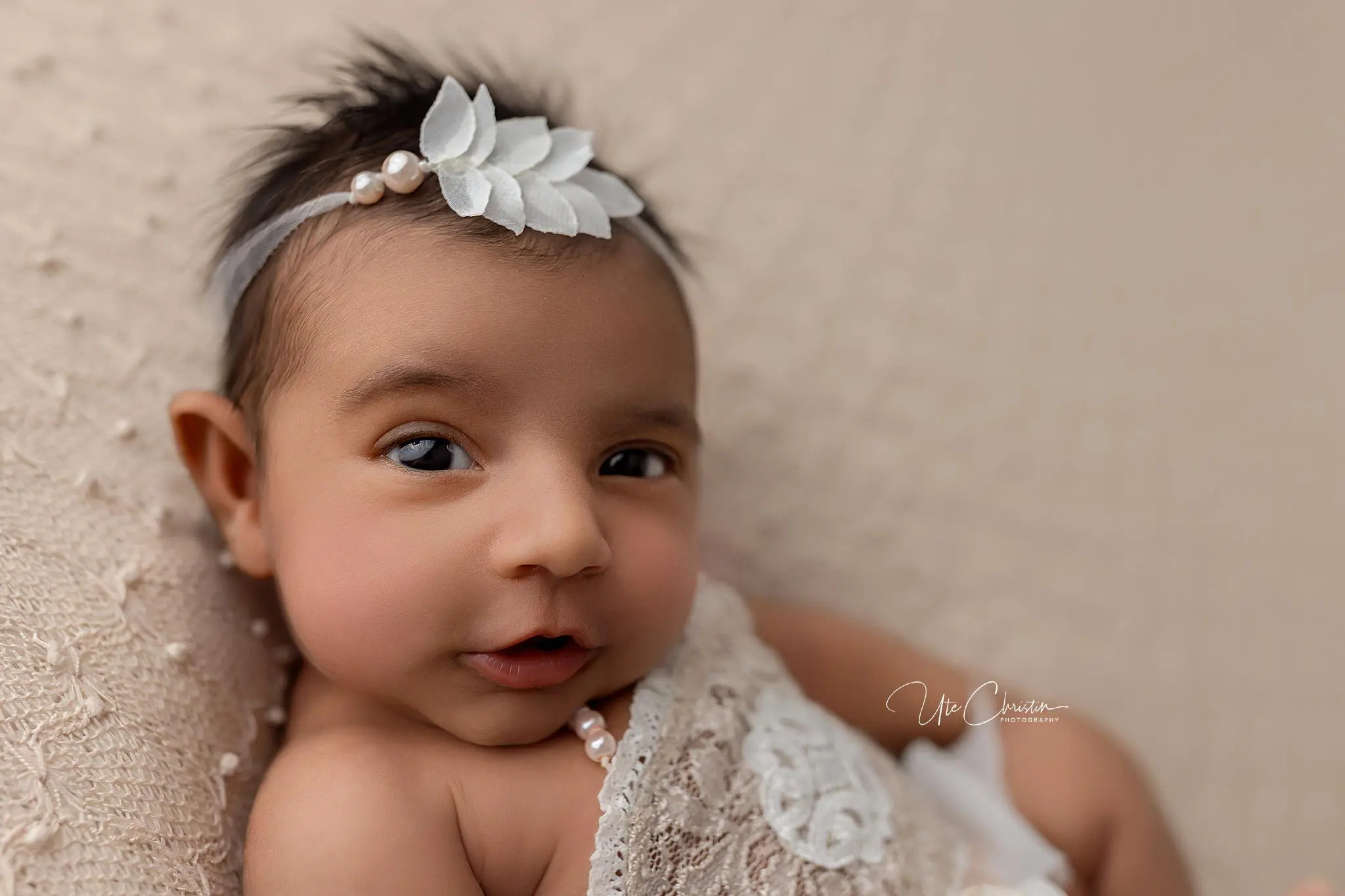 A close-up of a baby with dark hair wearing a white headband with leaves and pearl accents, lying on a textured beige blanket, wrapped in lace—perfect inspiration for Baby Shower Venues In Milford CT.