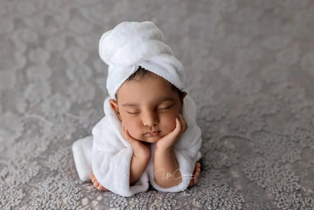 A baby wearing a white bathrobe and a towel wrapped around their head sleeps peacefully with their chin resting on their hands, lying on a soft, textured gray surface—perfect inspiration for Baby Shower Venues in Milford CT.