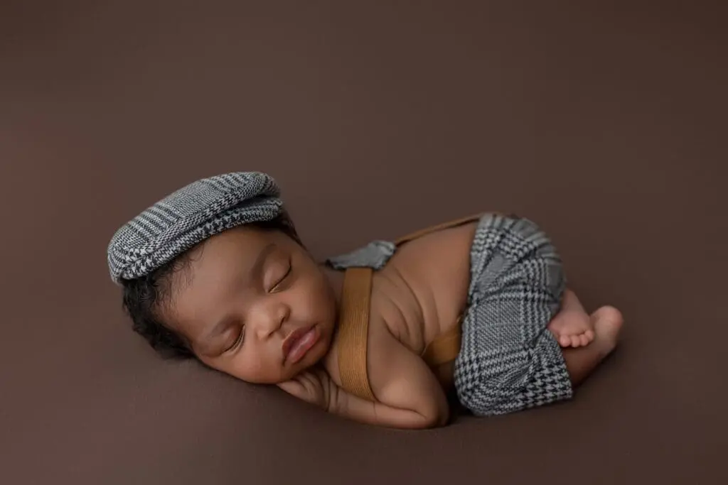 A sleeping newborn baby is curled up on a brown background, wearing a gray plaid cap and matching overalls. The serene scene, reminiscent of care from lactation consultants in CT, captures the baby positioned on their side with arms tucked under their head.