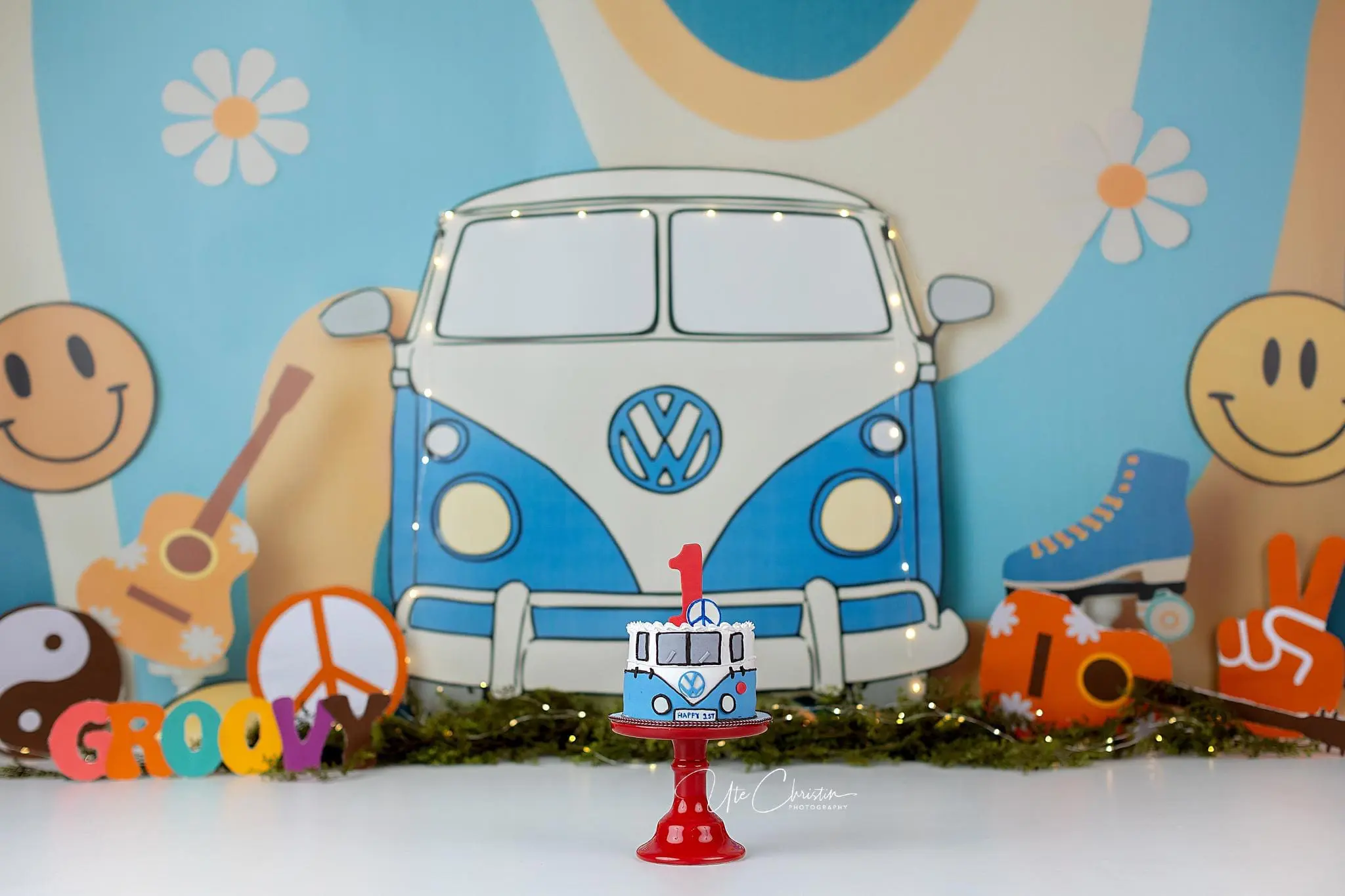 A blue and white retro van-themed cake with a red number 1 on top sits on a red stand. The backdrop features a colorful, groovy 1960s theme with a van illustration, smiley faces, flowers, and guitars.