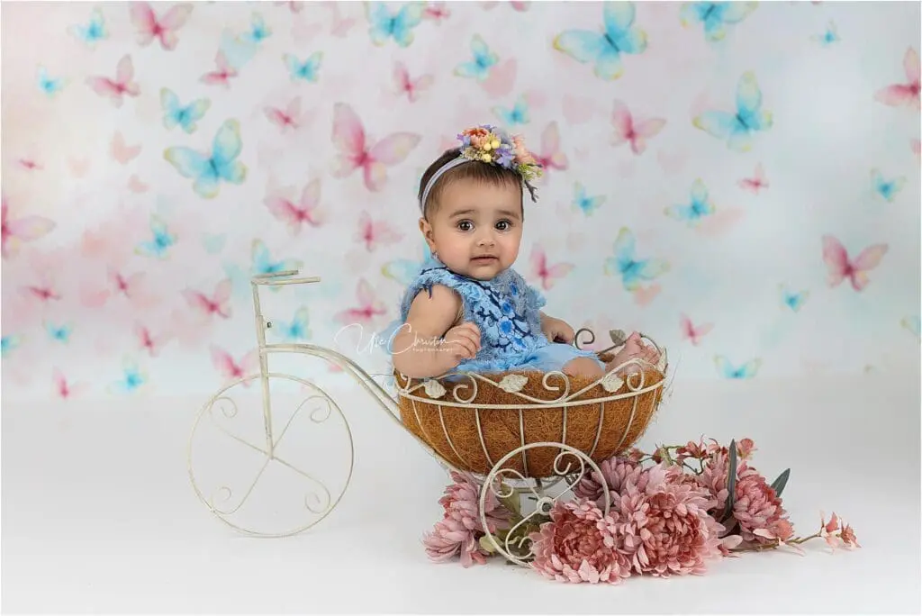 A baby wearing a blue dress and floral headband sits in a decorative bicycle basket, surrounded by pink flowers, with a pastel butterfly background—capturing the care and charm found with Nannies In Connecticut.