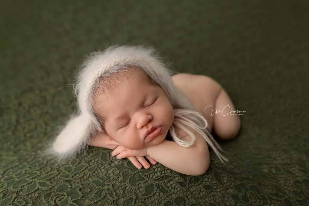 A newborn baby sleeps peacefully on a green lace blanket, wearing a soft, white knit bonnet with floppy ears, hands tucked under the chin—a serene moment often supported by caring postpartum doulas in CT.