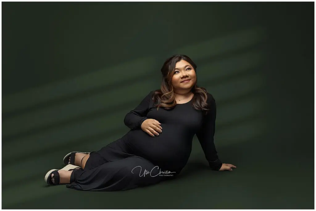 A pregnant woman in a long black dress sits on the floor against a dark green backdrop, smiling and resting one hand on her belly—perhaps relaxing after a Prenatal Massage In CT. Soft light creates diagonal shadows behind her.