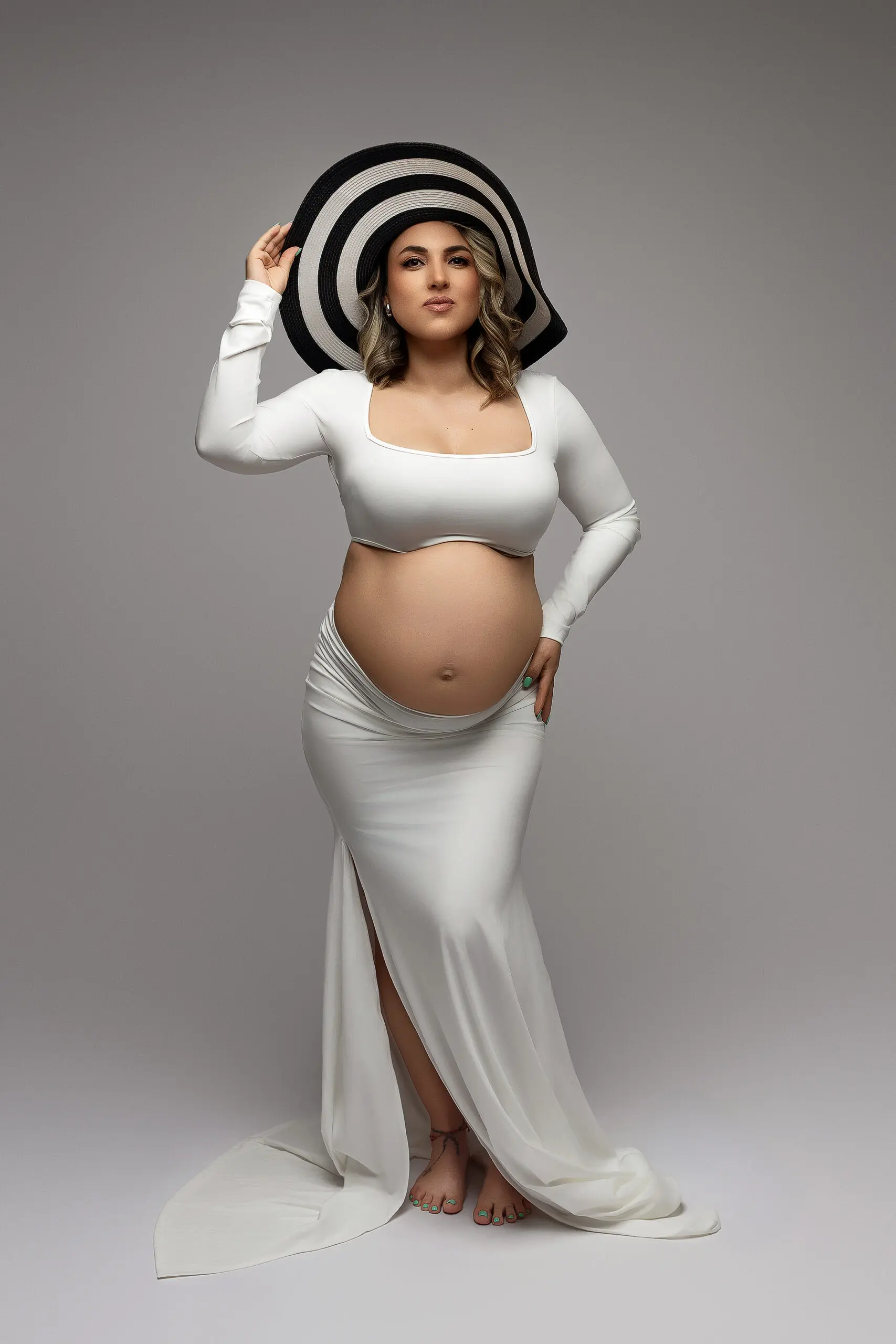 A pregnant woman in a white, long-sleeve crop top and skirt poses barefoot against a neutral background, holding a large black-and-white striped hat with one hand, revealing her baby bump during a maternity photoshoot in Connecticut.