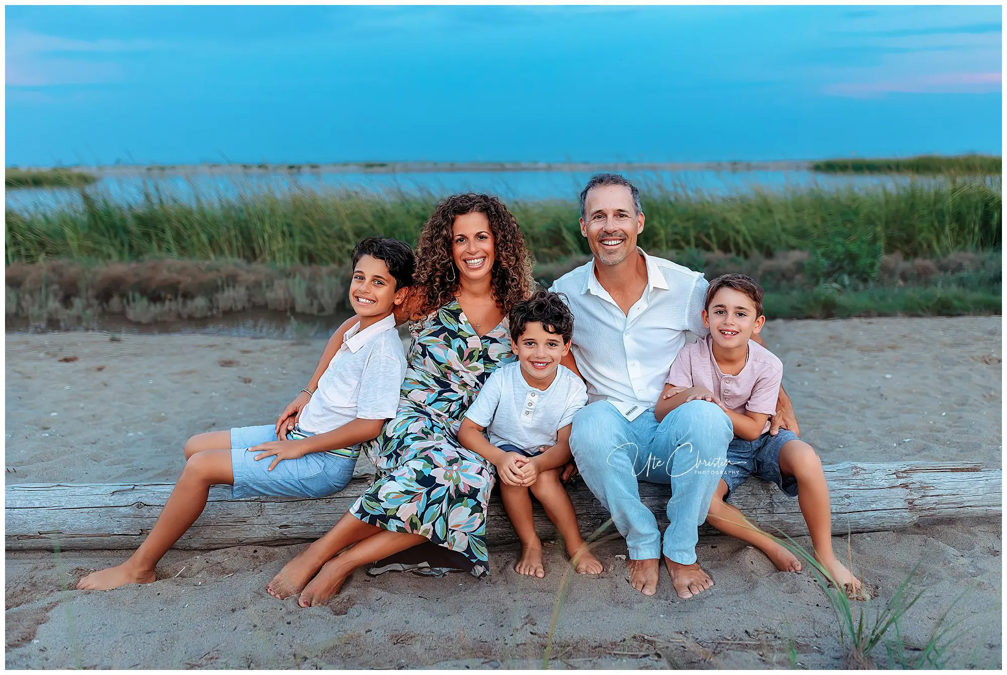 A family of five—two adults and three young boys—sit on a log at the beach, smiling at the camera. Tall grass and water are in the background under a blue sky at dusk.