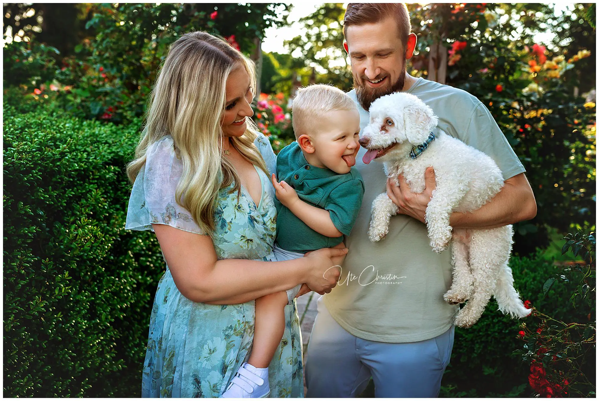 A smiling woman and man hold a laughing toddler and a fluffy white dog in a lush garden. The toddler reaches out to the dog, while all three adults and the dog look at each other happily.