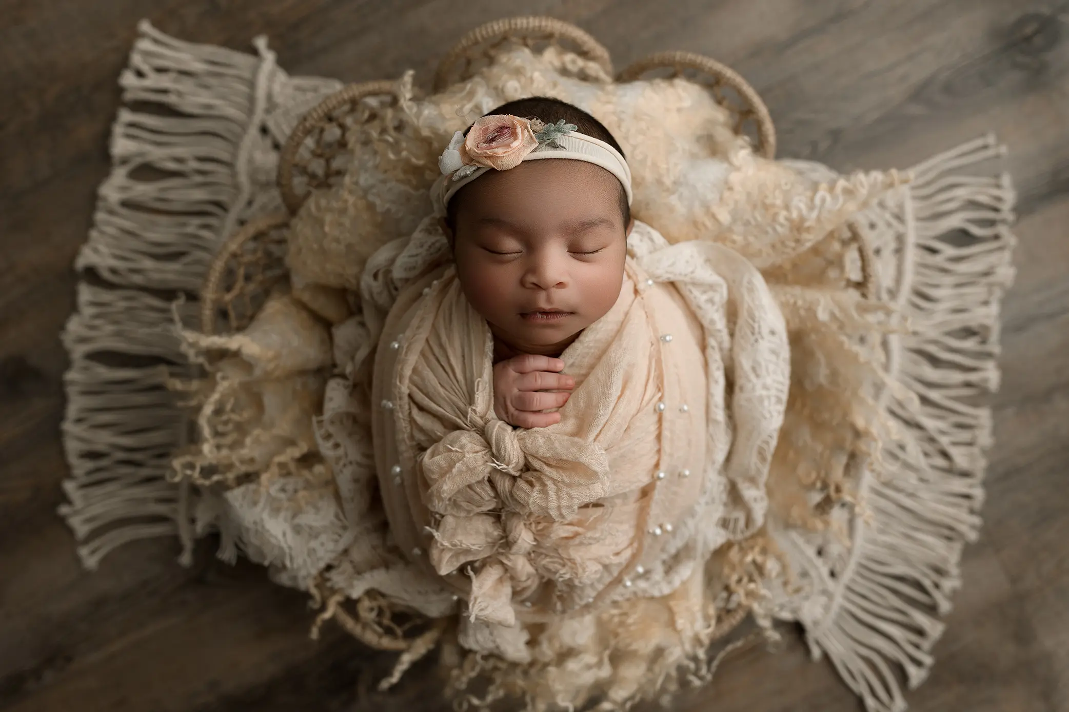 A newborn baby wrapped in a soft beige blanket, hands together, sleeps peacefully on a lacy, decorative fabric with fringe. The baby wears a floral headband and is lying on a wooden floor.