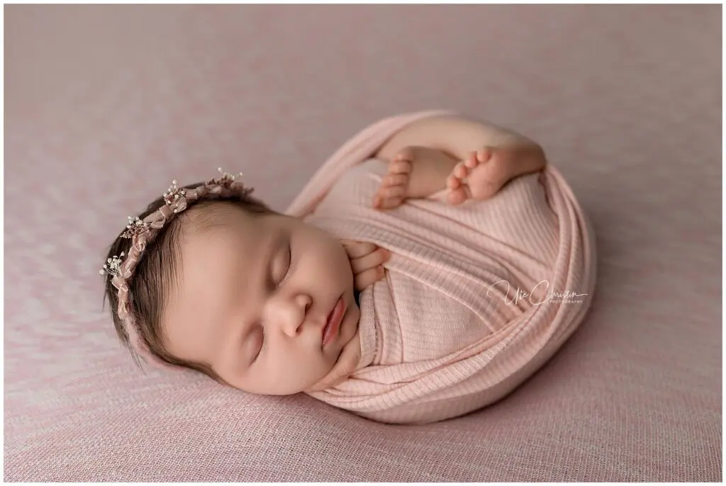 A newborn session in Milford captures a baby wrapped in a soft pink blanket, sleeping peacefully on a matching pink layer and wearing a delicate floral headband.