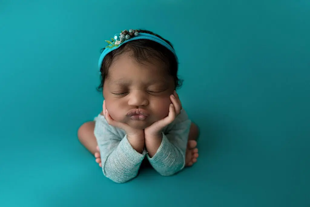 A newborn baby wearing a blue headband sits with elbows on knees, chin resting on hands, and eyes closed, against a teal background in a newborn studio near new haven connecticut.