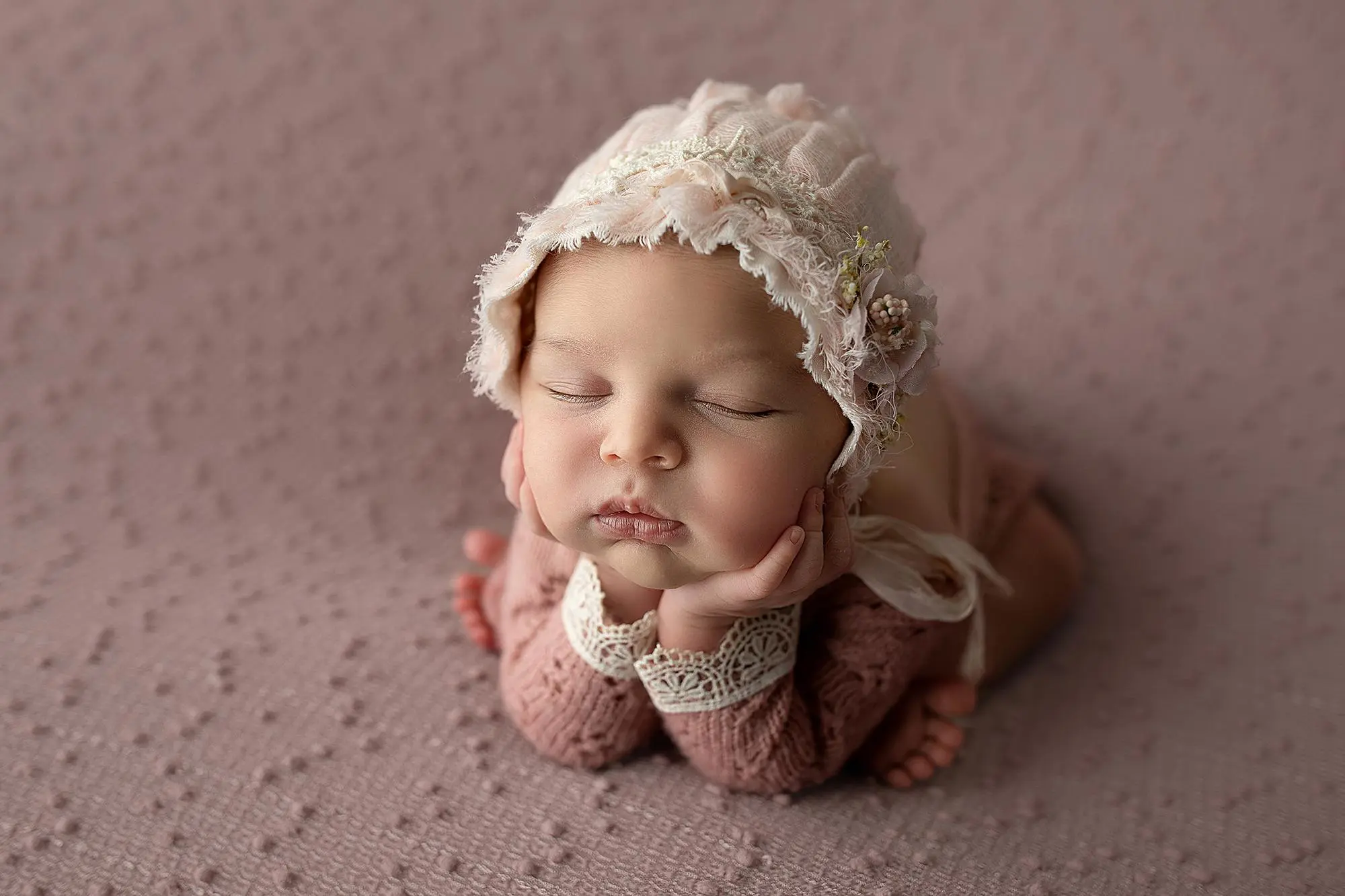 A newborn baby wearing a lace bonnet and a knitted outfit sleeps peacefully with their chin resting on their hands against a soft, textured pink blanket background during her new haven photography session.