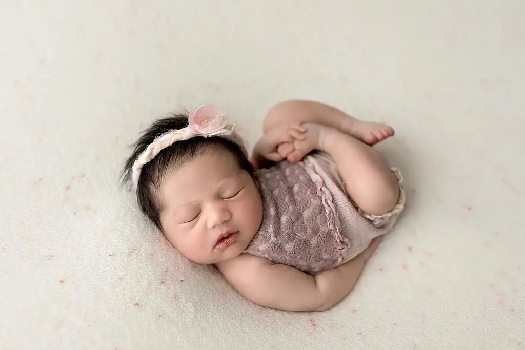 A newborn baby sleeps peacefully on a soft, light-colored blanket, wearing a lacy mauve outfit and a floral headband. The babys arms and legs are gently curled, and she has dark hair and closed eyes in New Haven.