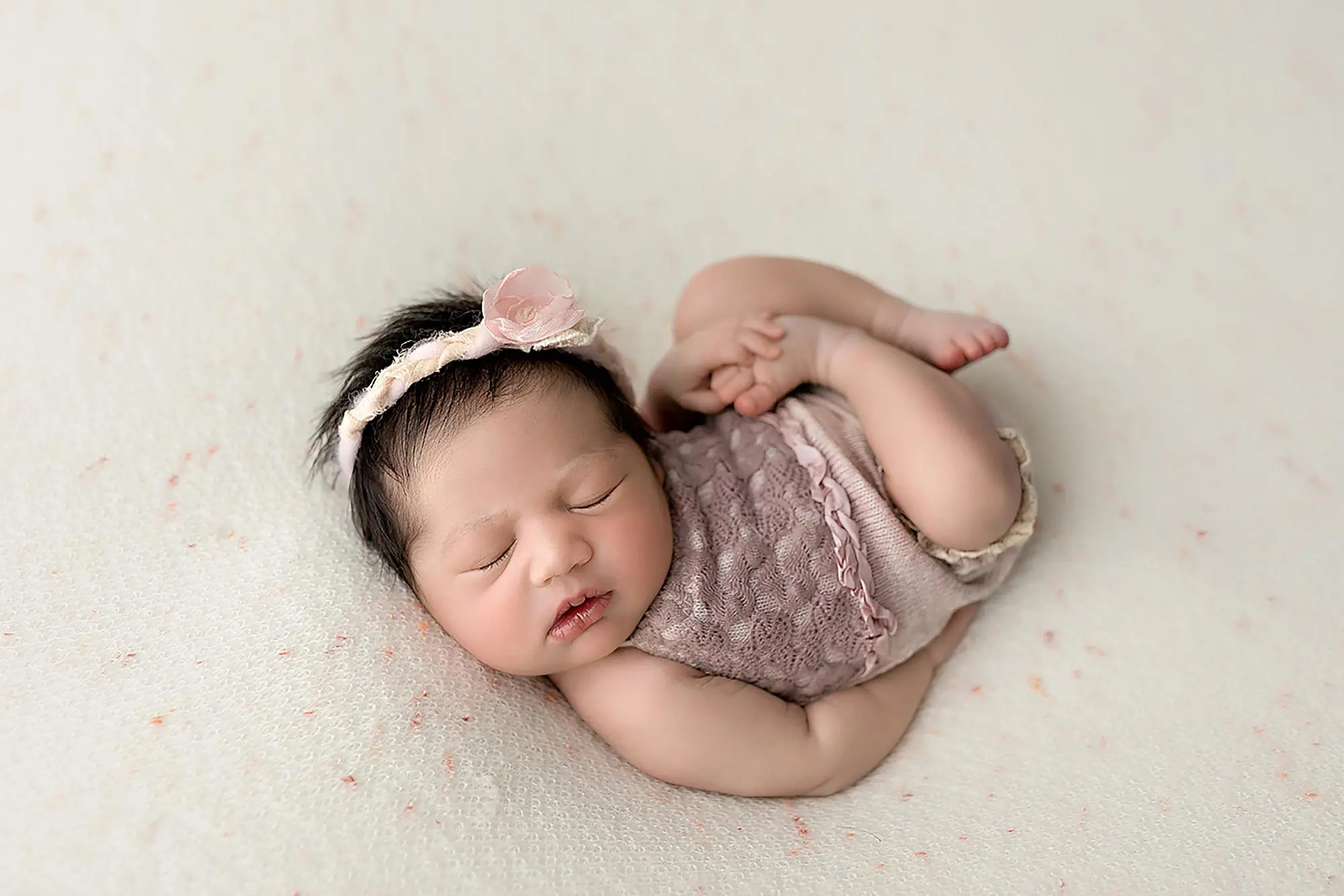 A newborn baby sleeps peacefully on a soft, light-colored blanket, wearing a lacy mauve outfit and a floral headband. The babys arms and legs are gently curled, and she has dark hair and closed eyes in New Haven.