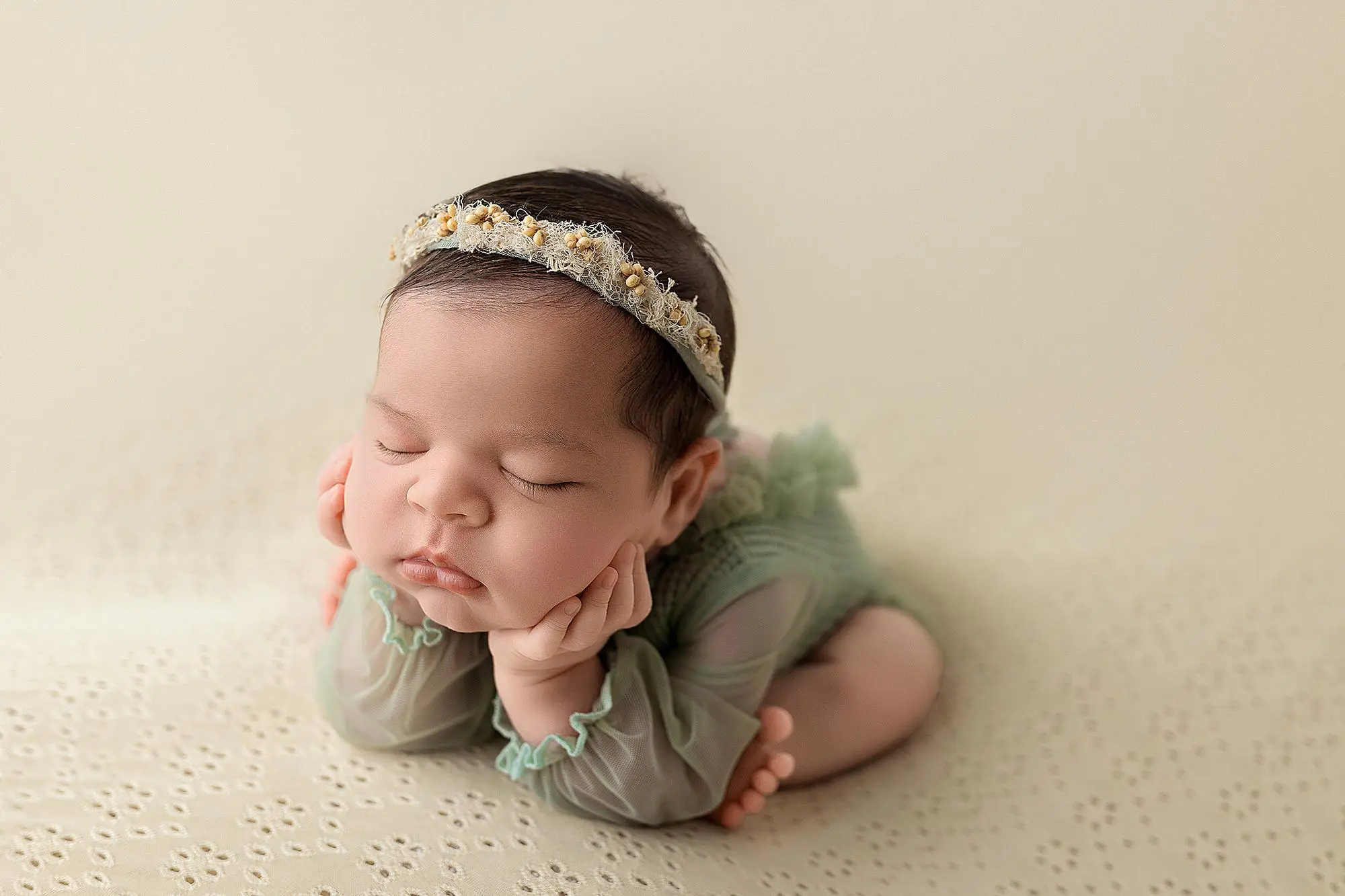 A newborn baby wearing a green outfit and a floral headband sleeps with their head resting on their hands, posed on a soft, cream-colored blanket during her newborn photos in new haven.