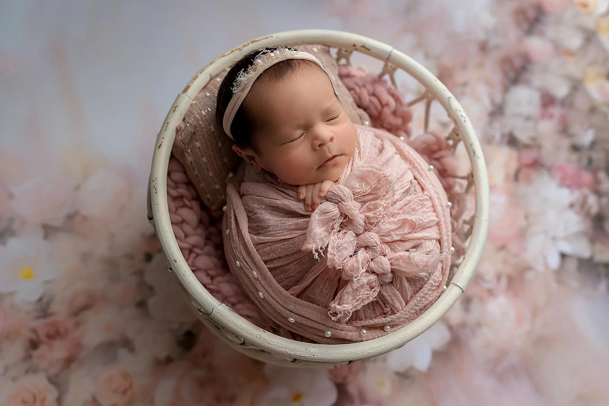 A newborn baby, wrapped in a soft pink blanket with a matching headband, sleeps peacefully in a round white basket. The background is decorated with soft, pastel-colored flowers for her newborn photos in new haven.