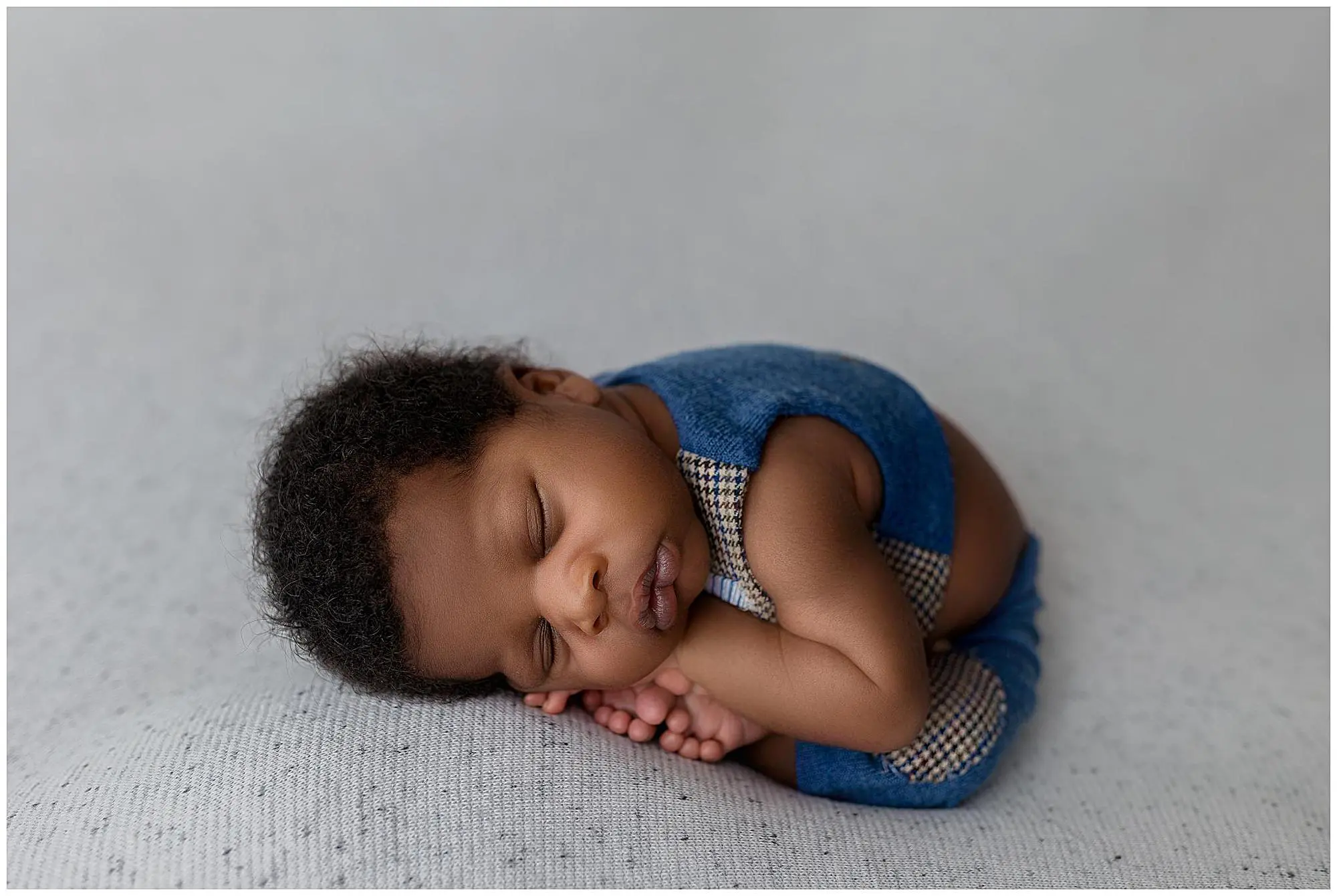 A newborn baby wearing a blue outfit sleeps peacefully on a soft, light gray surface, curled up with hands tucked under their cheek and feet drawn close to their body during their newborn photo session.