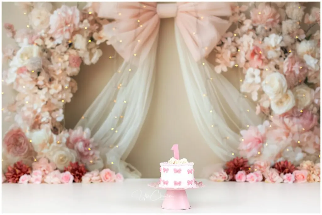 A small pink and white cake with a number 1 topper sits on a stand. Behind it is a backdrop of pink, cream, and white flowers, sheer fabric, a large bow, and twinkling fairy lights.