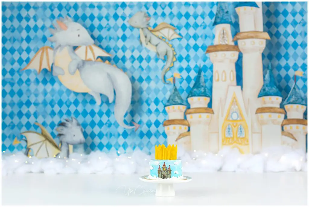 A small blue cake with a castle and yellow crown decoration sits on a white stand. The background features illustrated dragons, a castle, and a blue diamond pattern, with white clouds at the bottom.