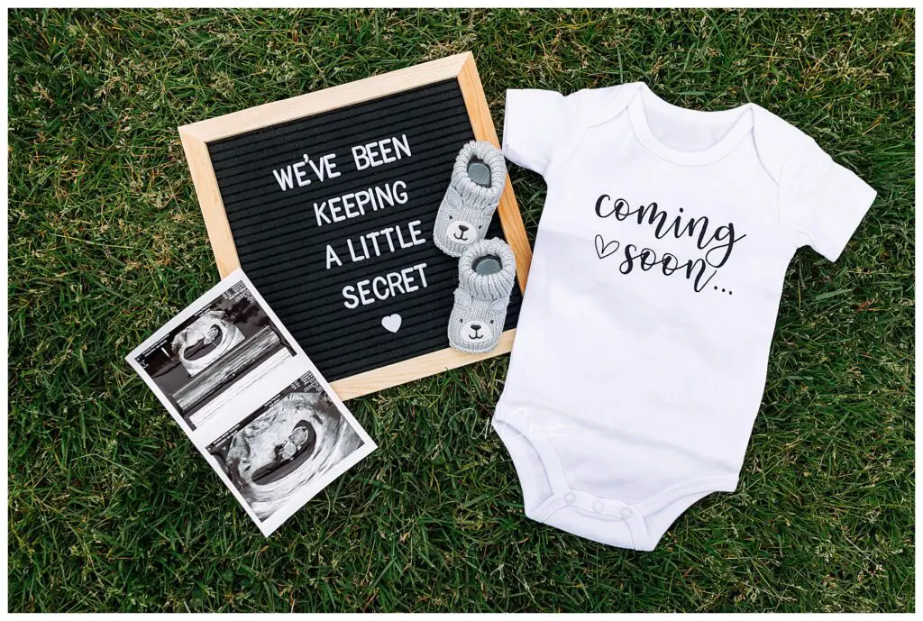 A flat lay on grass showing a letterboard with “We’ve been keeping a little secret,” baby sonogram photos, gray baby booties, and a white onesie with “coming soon...” written on it.