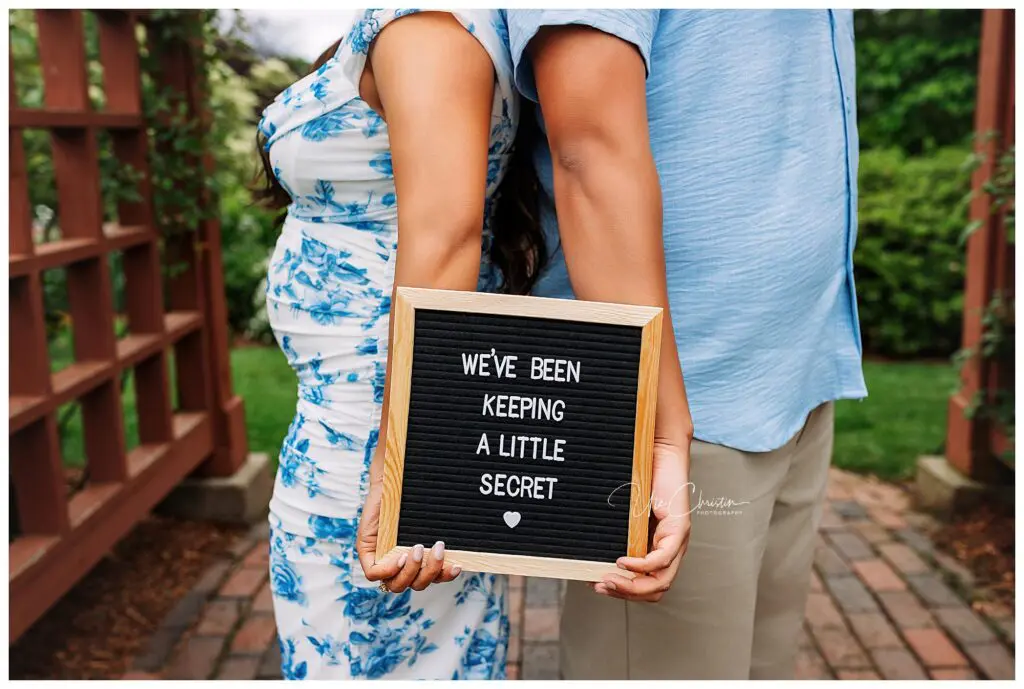 A couple stands back to back outdoors, each holding a letterboard that reads “We’ve been keeping a little secret” with a small heart below. The woman, in a blue floral dress, smiles—celebrating their journey with help from Connecticut Fertility Clinic.