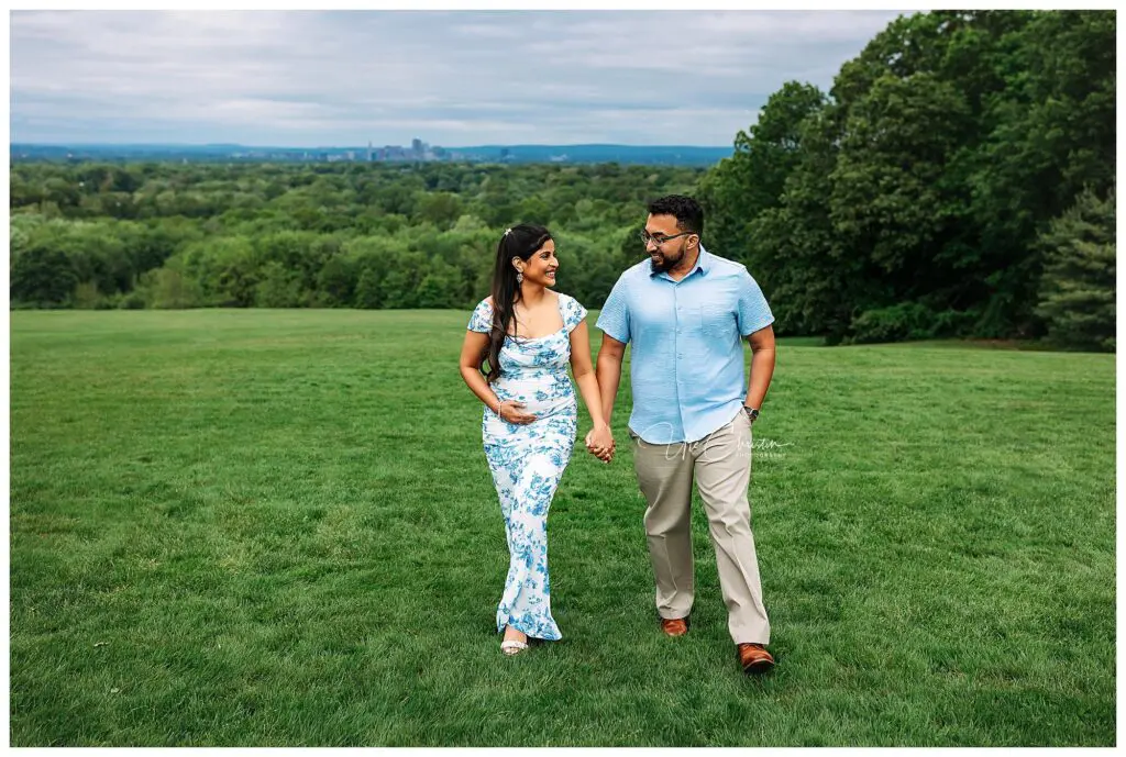 A couple holding hands and smiling at each other while walking on a grassy field, with green trees and a distant city skyline in the background. The woman is wearing a blue and white dress, and the man is in a blue shirt and beige pants.