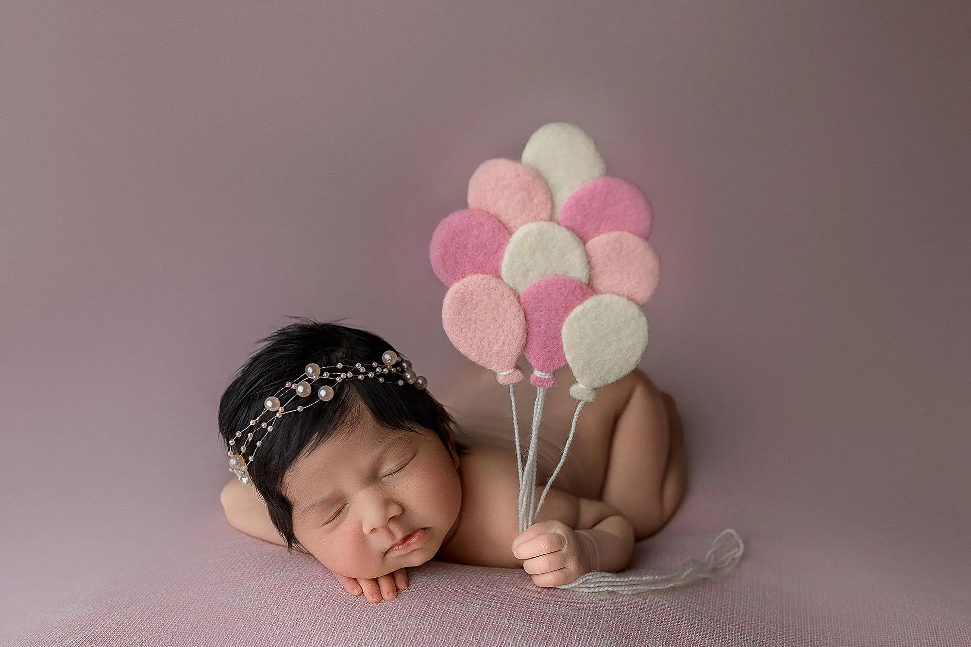 A sleeping newborn baby with a pearl headband lies on a soft pink surface, holding a bunch of pink and white felt balloon props.