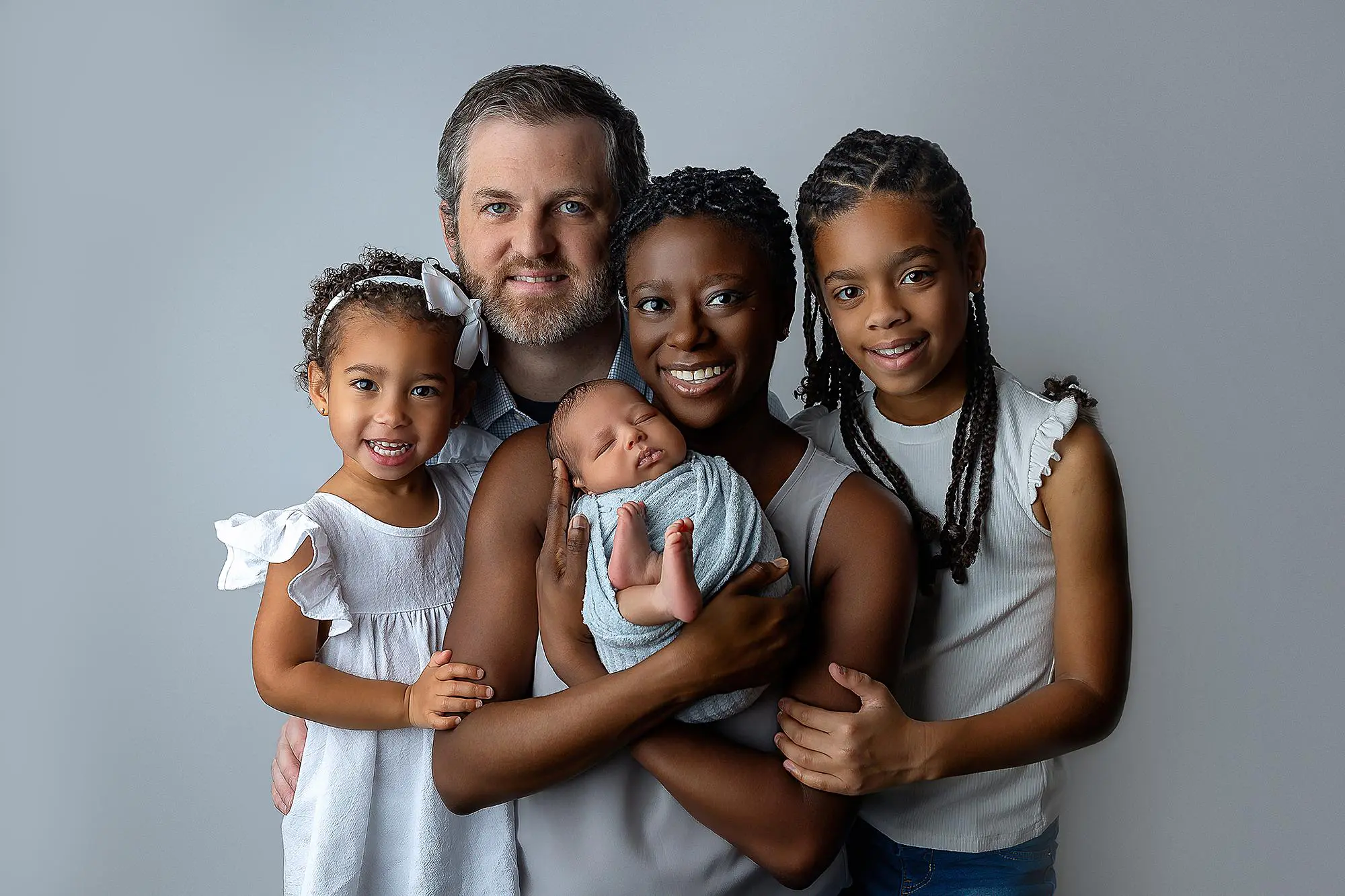 A smiling family of five poses against a light gray background. The parents hold a newborn baby, while their two daughters, both smiling, stand on either side. Everyone is dressed in light-colored clothing during their new haven newborn photo shoot.