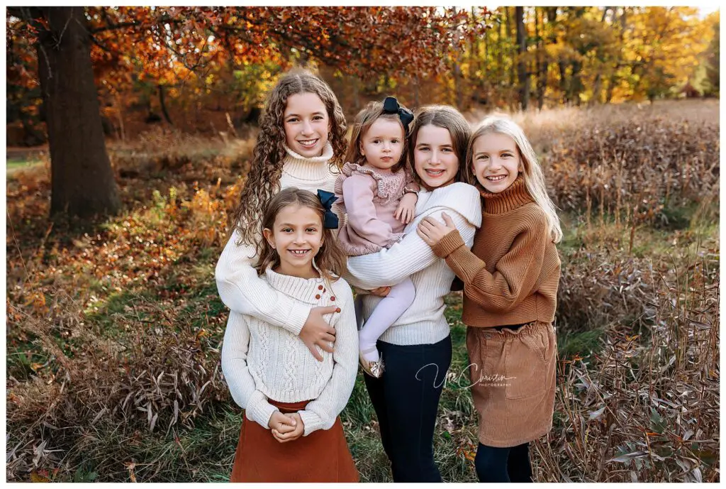 Five girls stand together outdoors in a field with autumn leaves and trees. They are smiling, wearing sweaters and skirts or dresses in fall colors, and one girl is holding a toddler in a pink dress with a black bow.