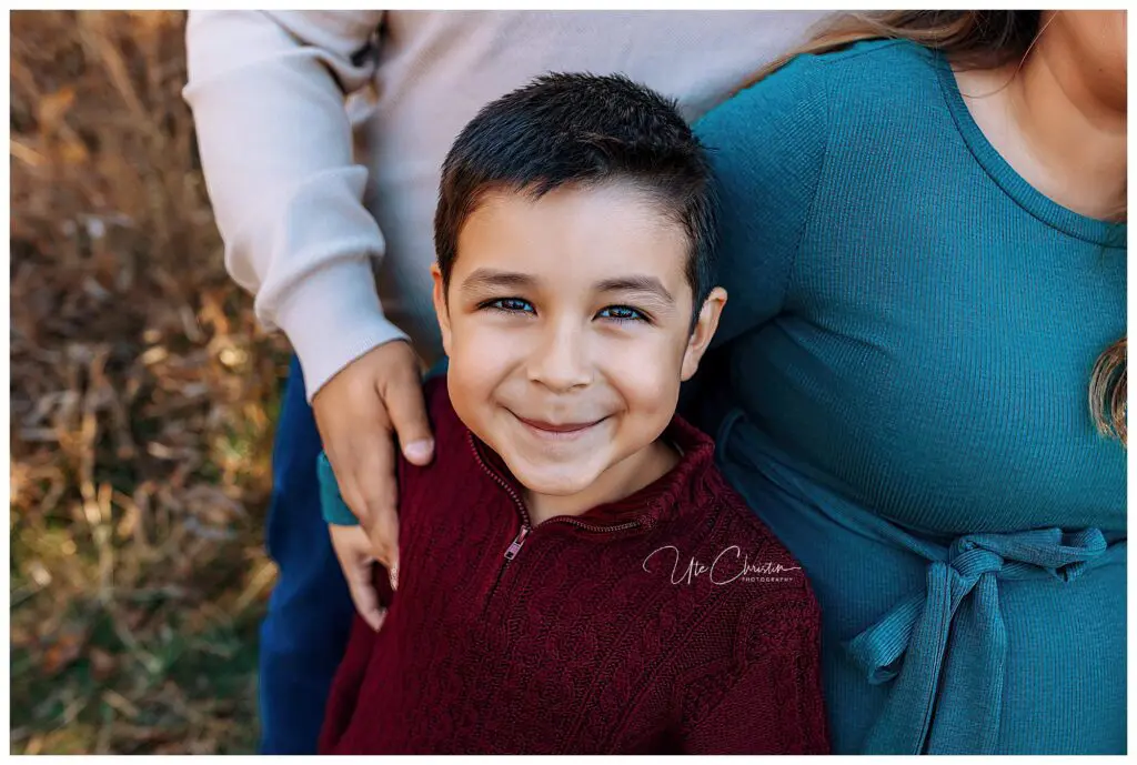 A young boy with short dark hair smiles at the camera, wearing a maroon sweater. He stands in front of two adults, one with an arm around him and the other wearing a teal dress, possibly his family, outdoors.
