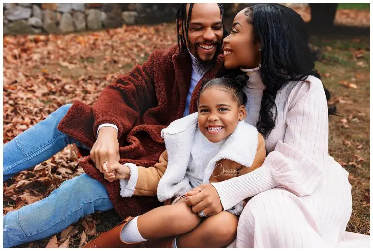 A family sits together on fallen leaves, smiling warmly. A man and woman, dressed in cozy autumn clothes, embrace a young girl wearing a brown jacket and boots, who sits in front and grins at the camera.