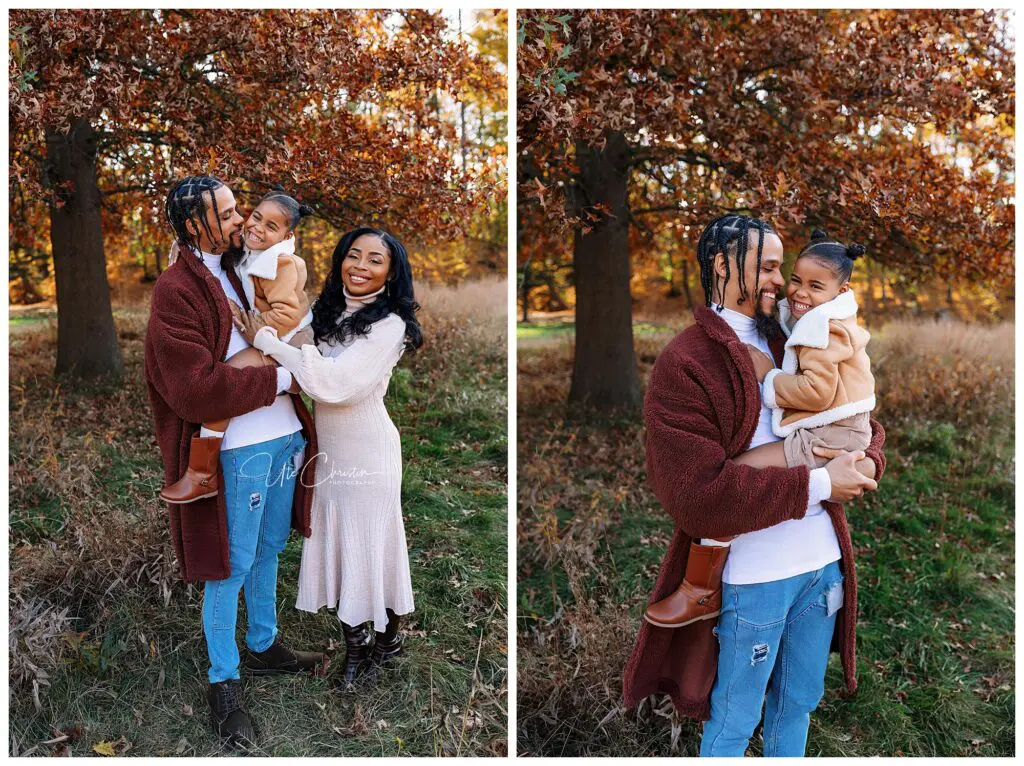 A smiling family of three poses outdoors in autumn. The father holds their daughter while the mother stands beside them. Fall foliage with orange leaves fills the background. They all wear cozy, casual clothes.