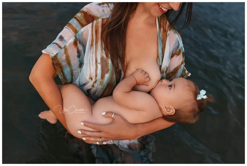 A woman stands in water breastfeeding a baby with a blue bow in her hair. The woman wears a sheer, colorful cover-up and smiles while holding the baby in her arms.