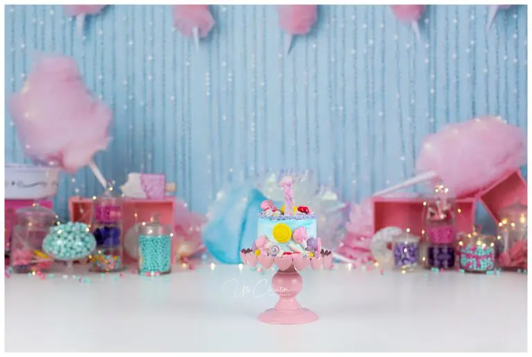 A pastel-colored cake topped with candy and a pink number one sits on a pink stand. The background features jars of colorful sweets, cotton candy, and twinkling fairy lights against a light blue backdrop.