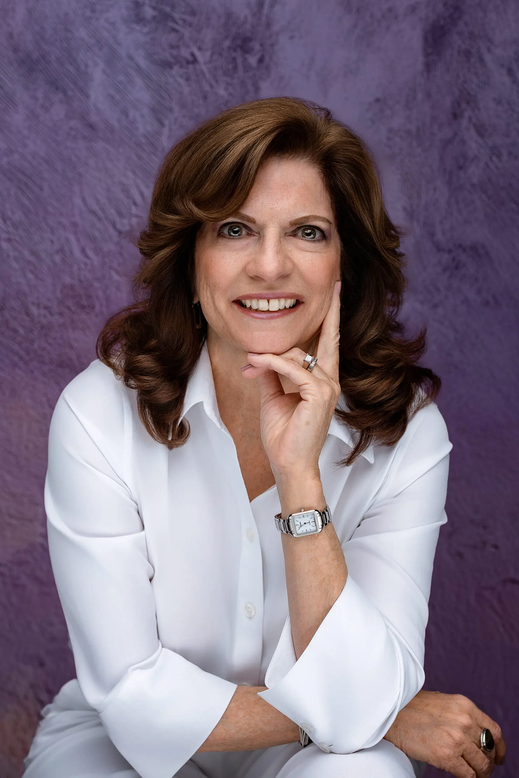 A woman with wavy brown hair, wearing a white shirt, silver watch, and ring, smiles softly while resting her chin on her hand, seated in front of a textured purple background for her CT headshot.