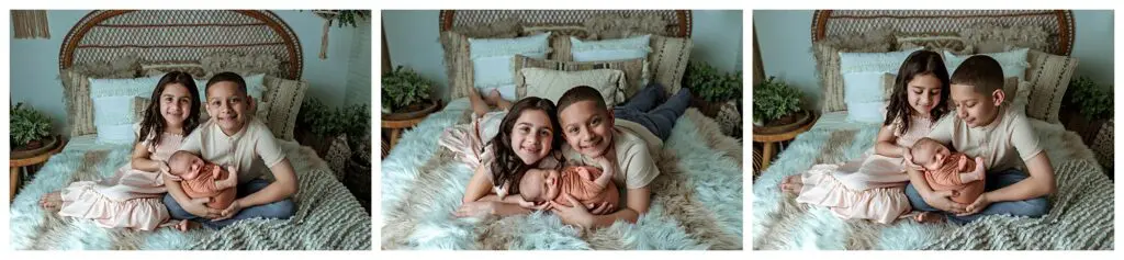 Three children sit and lie together on a cozy bed with fluffy white bedding; the older girl and boy cradle a newborn wrapped in peach fabric, all looking happy and close.