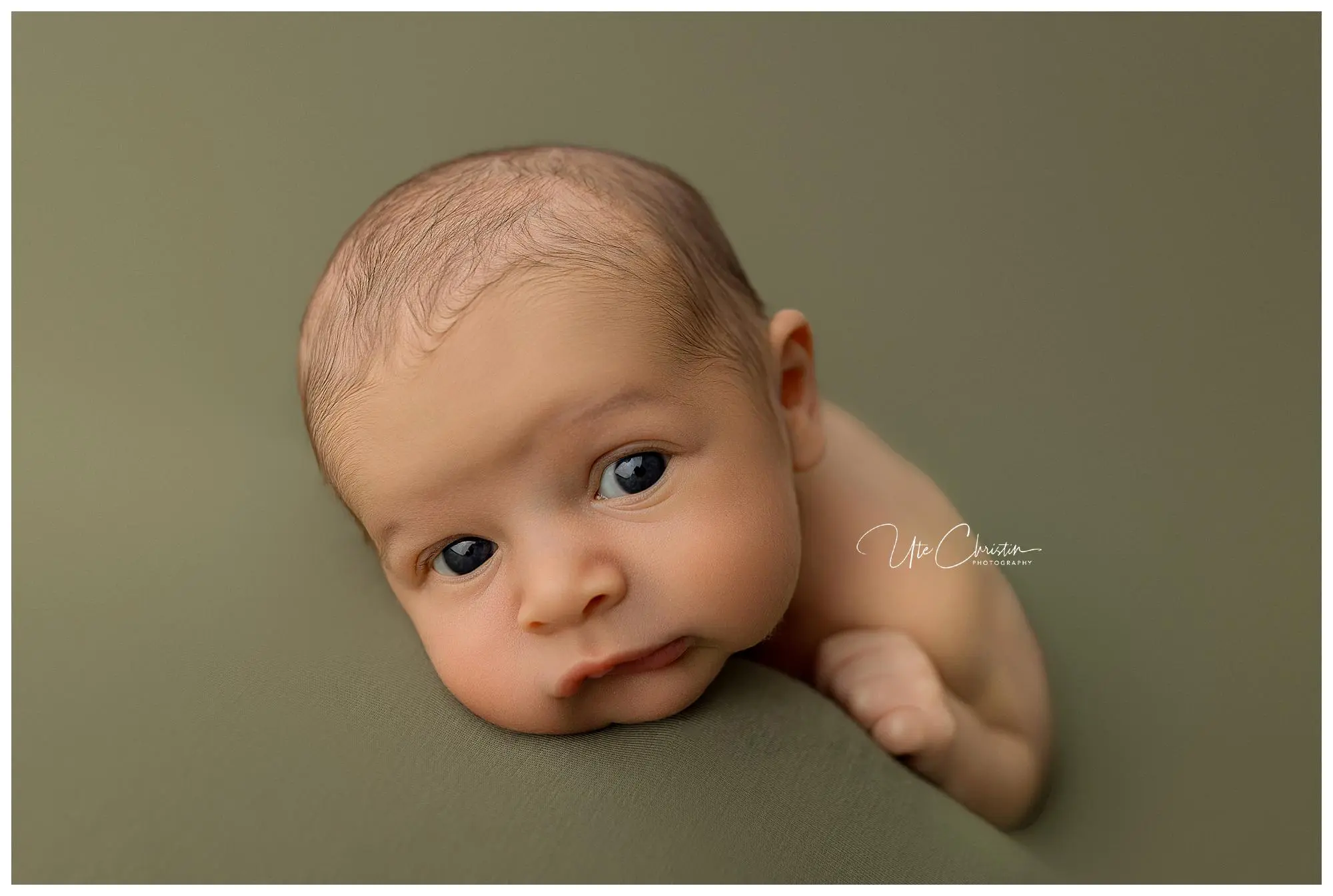 A newborn baby with big, curious eyes rests on a soft, olive-green surface, looking directly at the camera with a calm expression.