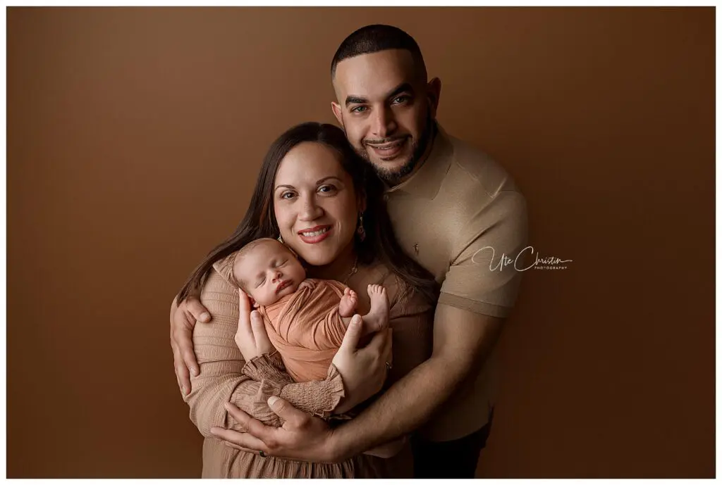 A smiling couple poses in front of a brown background, with the woman holding a sleeping newborn dressed in a peach outfit. The man stands behind the woman, embracing both her and the baby lovingly.