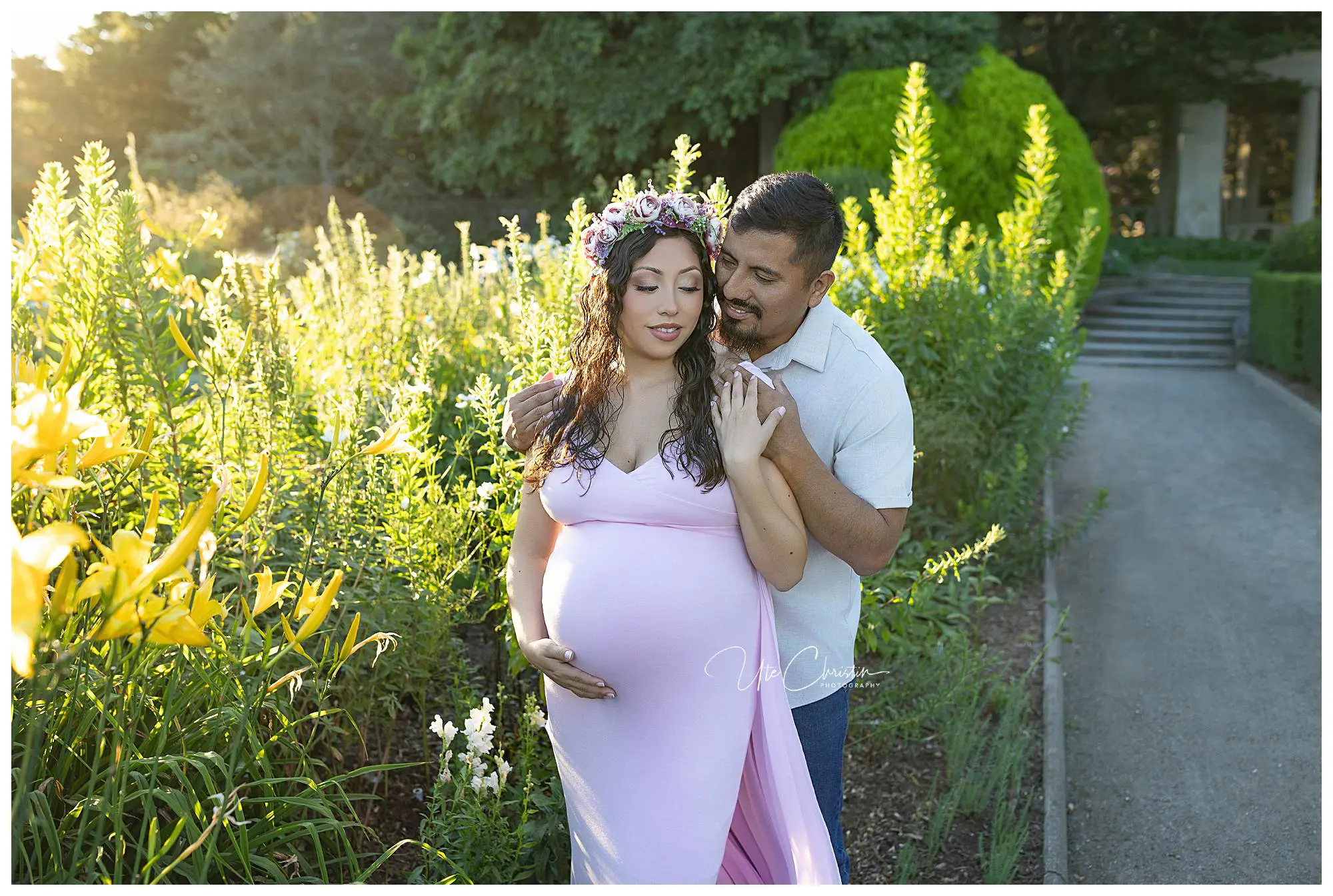 A couple poses outdoors in a garden; the woman, wearing a pink dress and floral crown, cradles her pregnant belly while the man stands behind her, gently holding her shoulders. Sunlight filters through the flowers around them.