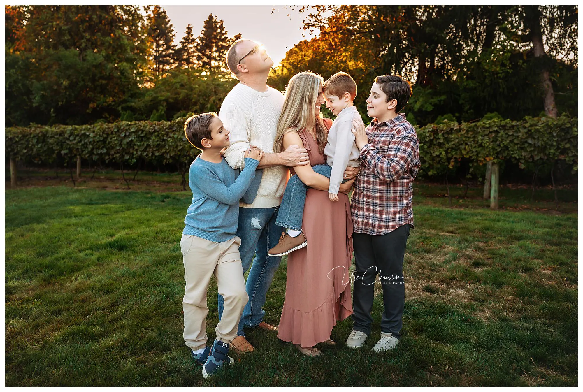 A family of five stands together outdoors on green grass at sunset, smiling and embracing. The parents are in the center, with three boys around them, surrounded by trees and a warm, glowing sky in the background.