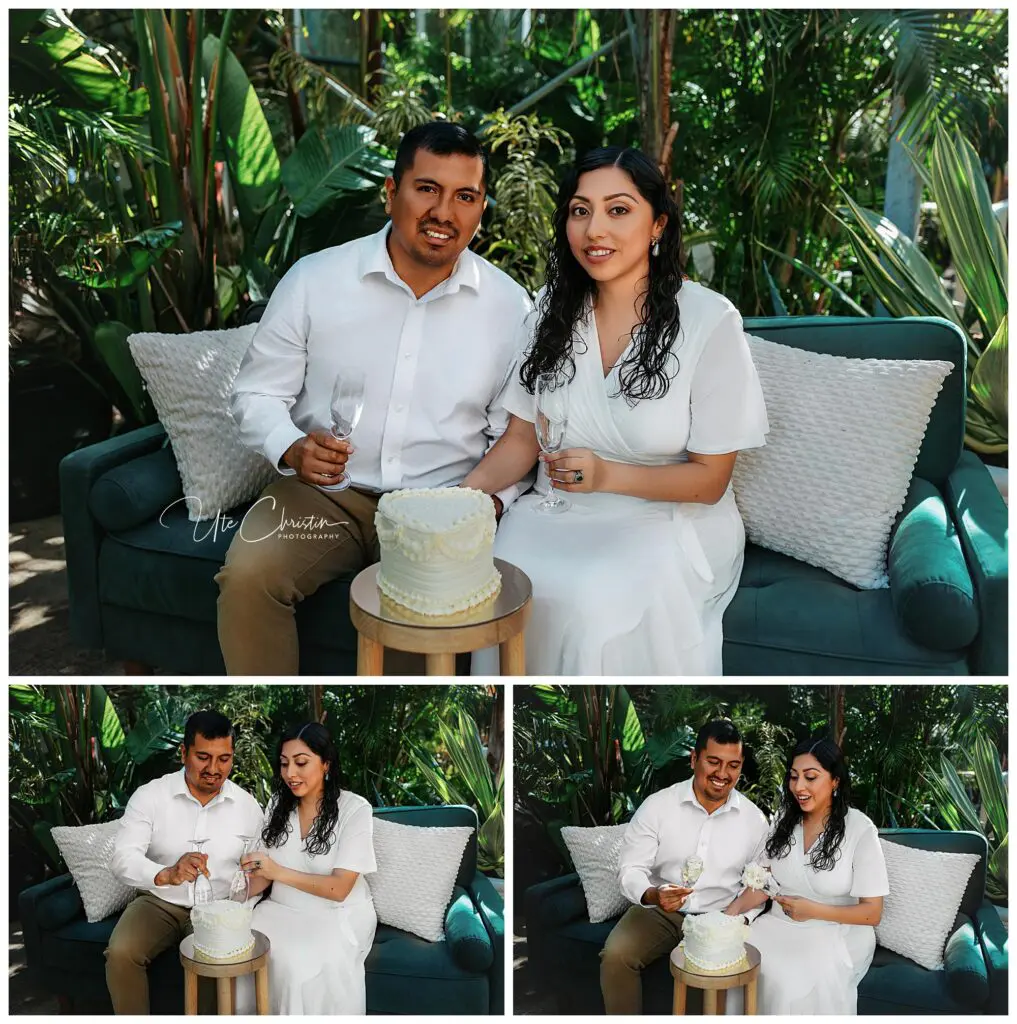 A couple dressed in white sits on a green couch outdoors, holding champagne glasses and posing with a small white cake on a table. They smile while preparing to cut the cake in a lush, garden setting.