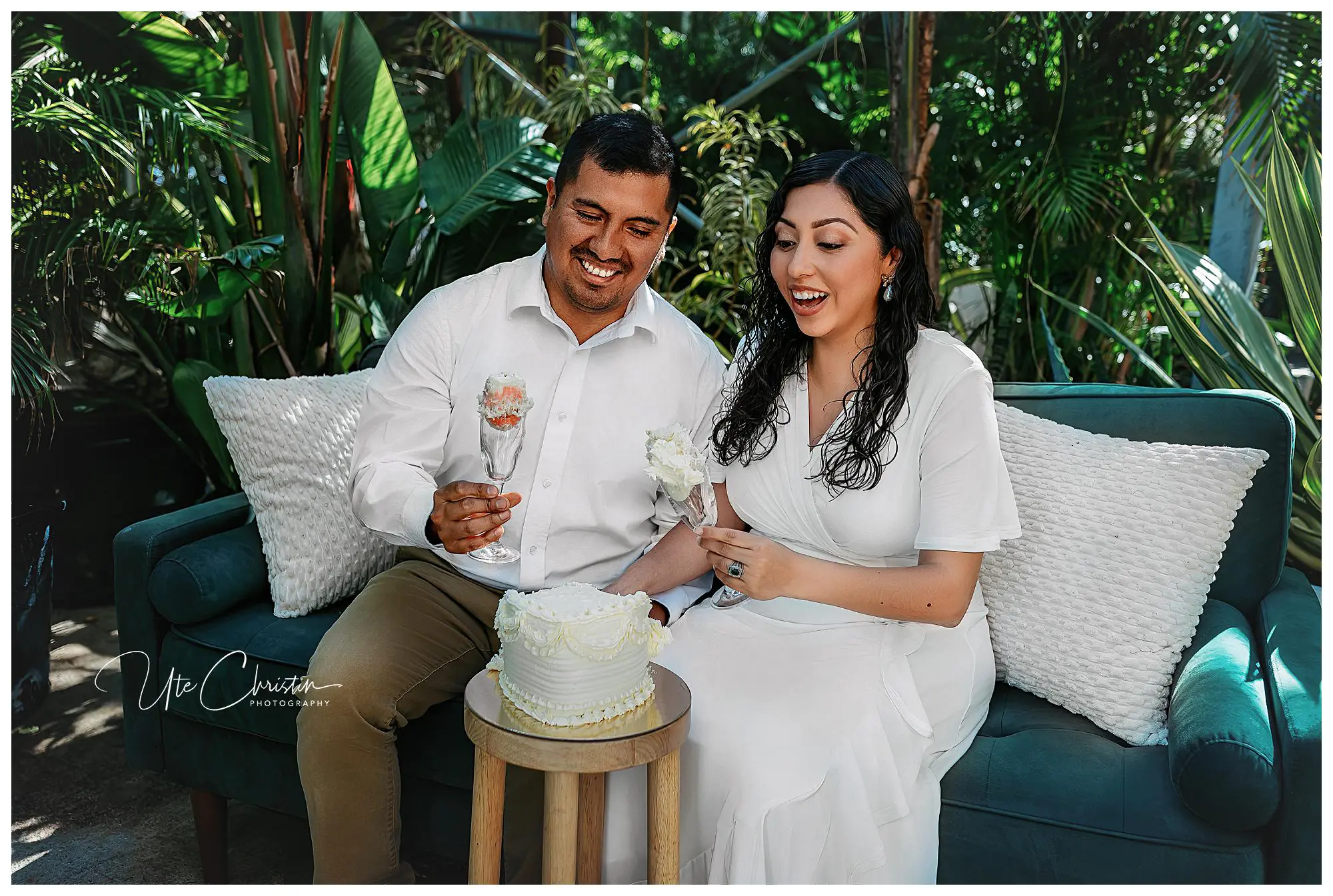 A smiling couple in white attire sit on a green couch outdoors, holding cake and glasses, with a small white-frosted cake on a table in front of them. Lush greenery surrounds them, creating a festive atmosphere.