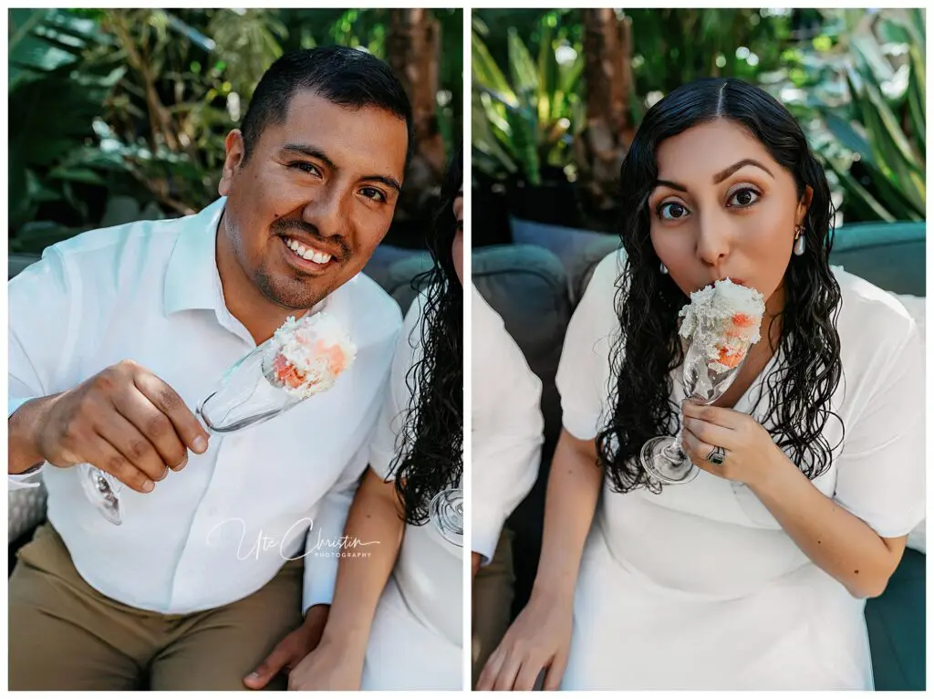 A man and a woman, both in white outfits, sit outdoors holding dessert-filled champagne glasses, smiling and enjoying their treats. Greenery surrounds them in the background.