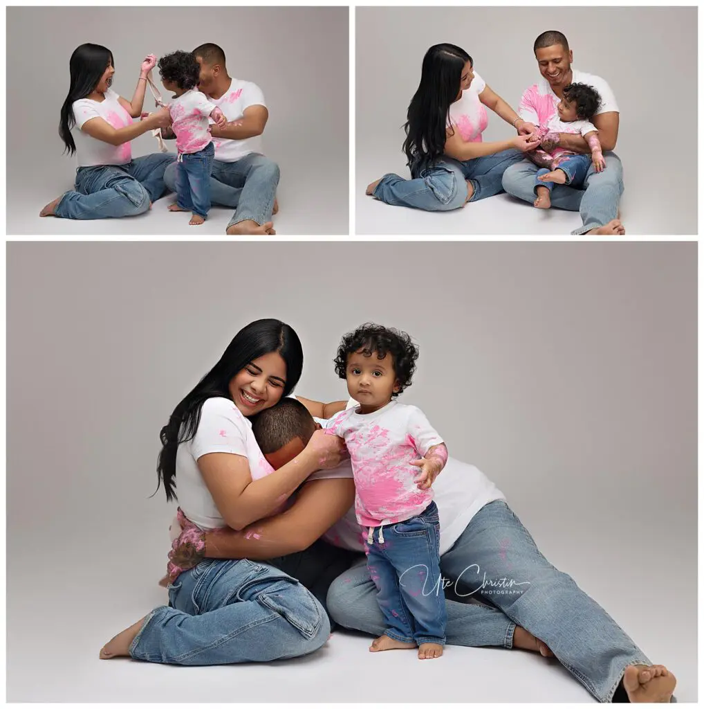 A family of three, wearing matching white shirts and jeans, playfully interact in a studio. The child has pink paint on their shirt, and the parents smile and hug, creating a joyful, candid moment together.
