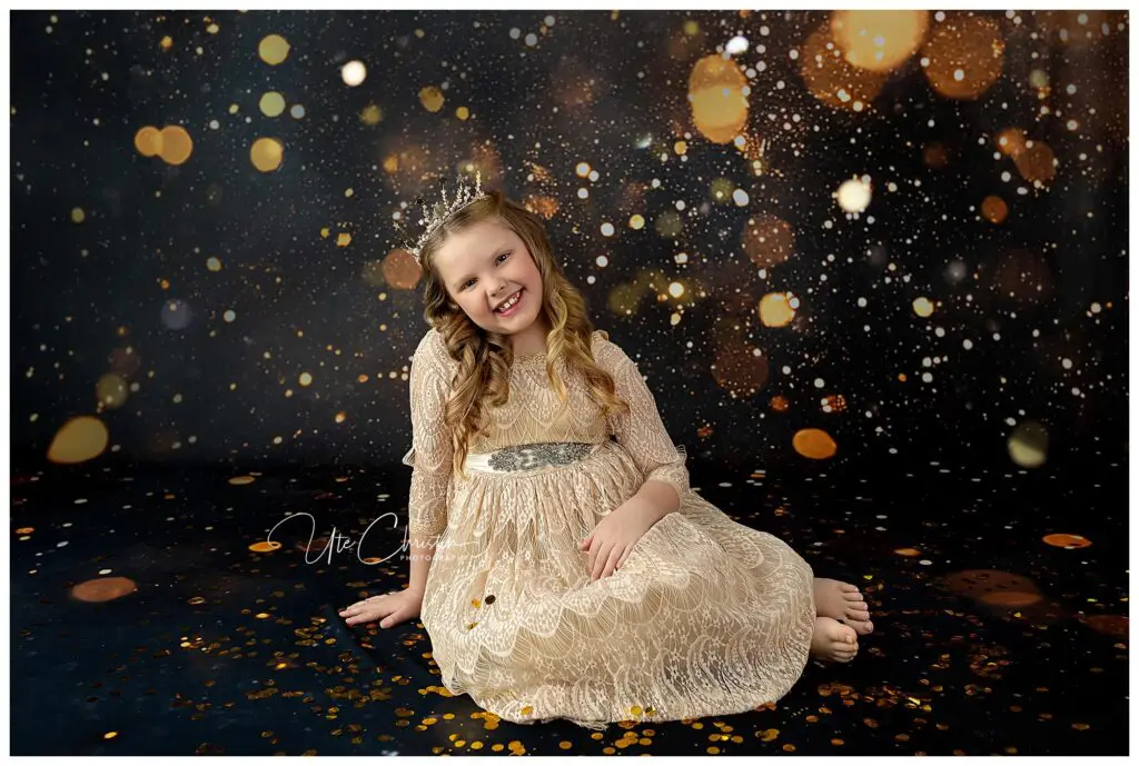 A young girl in a cream lace dress and tiara sits barefoot on the floor, smiling against a dark background with golden bokeh and sparkling light effects, creating a magical, festive atmosphere.
