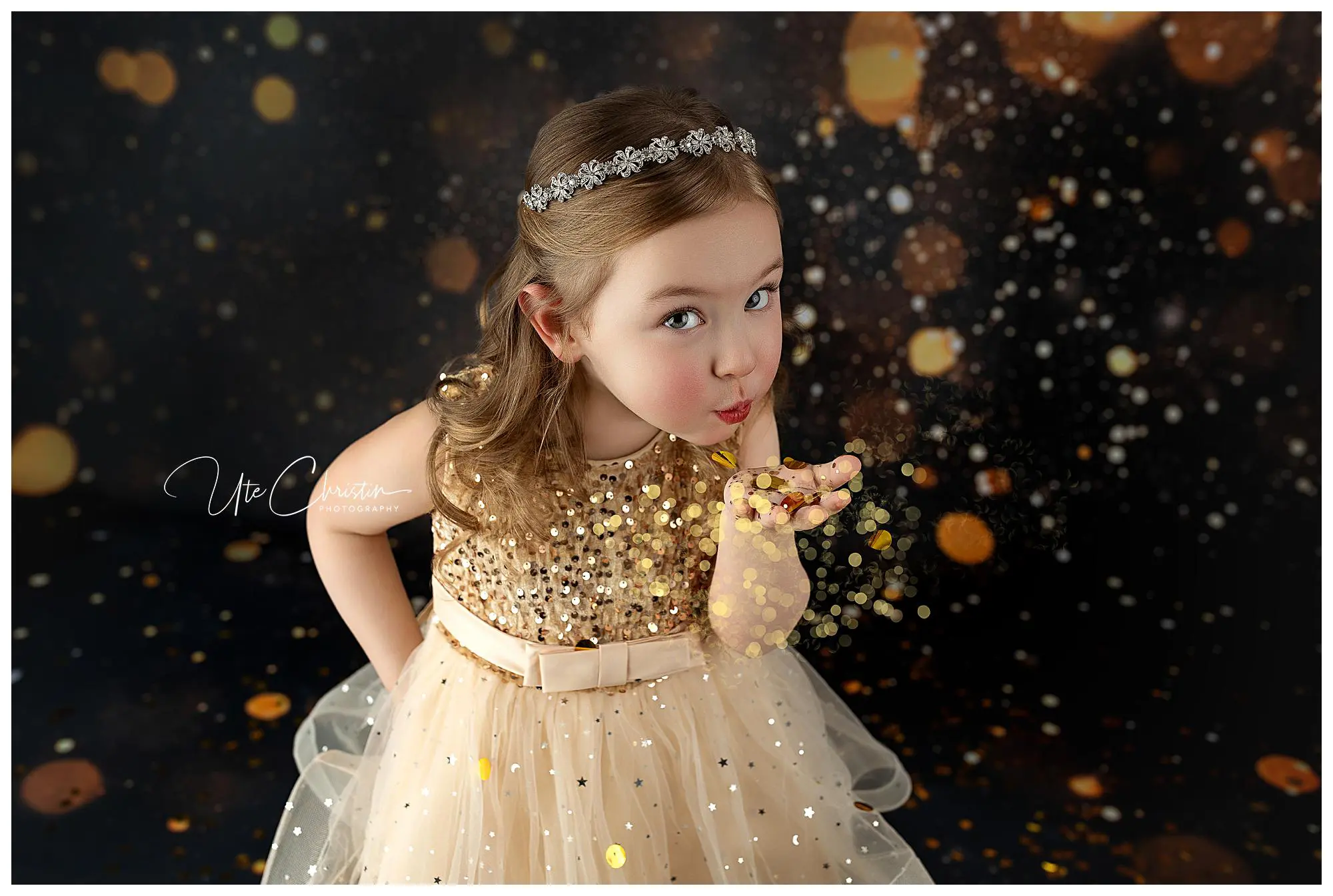 A young girl in a gold sequin dress and jeweled headband blows glitter from her hand against a dark background with sparkling gold light effects. She looks playful and festive.