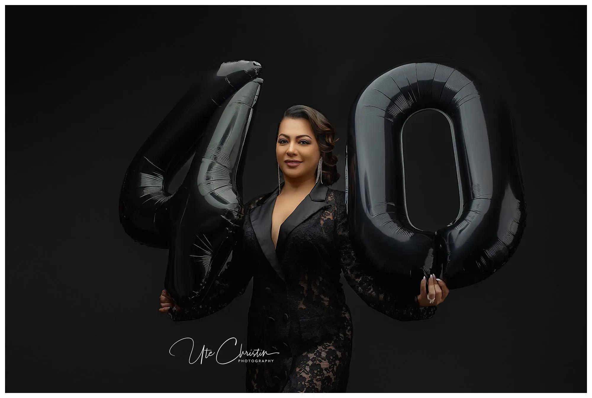 A woman in a black lace outfit holds large black balloons shaped as the number 40, standing against a dark background and smiling confidently at the camera.