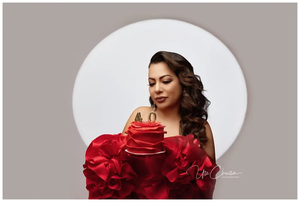 A woman with styled hair and makeup holds a red rose-shaped cake with gold 40 candles against a white circle background, wearing a dress adorned with large red ruffles.