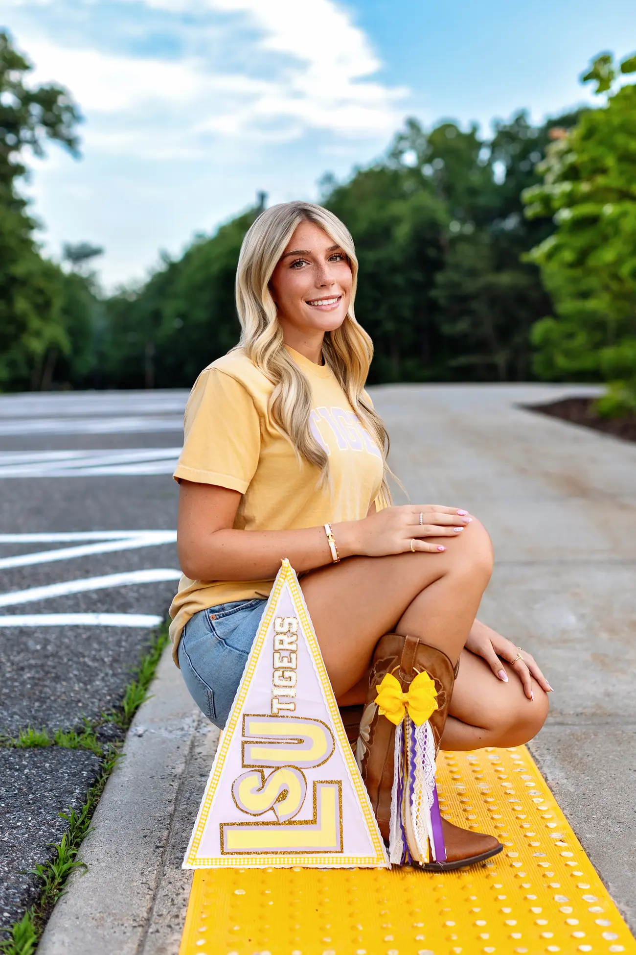 A young woman with long blonde hair, wearing an LSU shirt, denim shorts, and brown boots with yellow tassels, kneels on a yellow curb next to an LSU Tigers pennant. Trees and a blue sky are in the background.